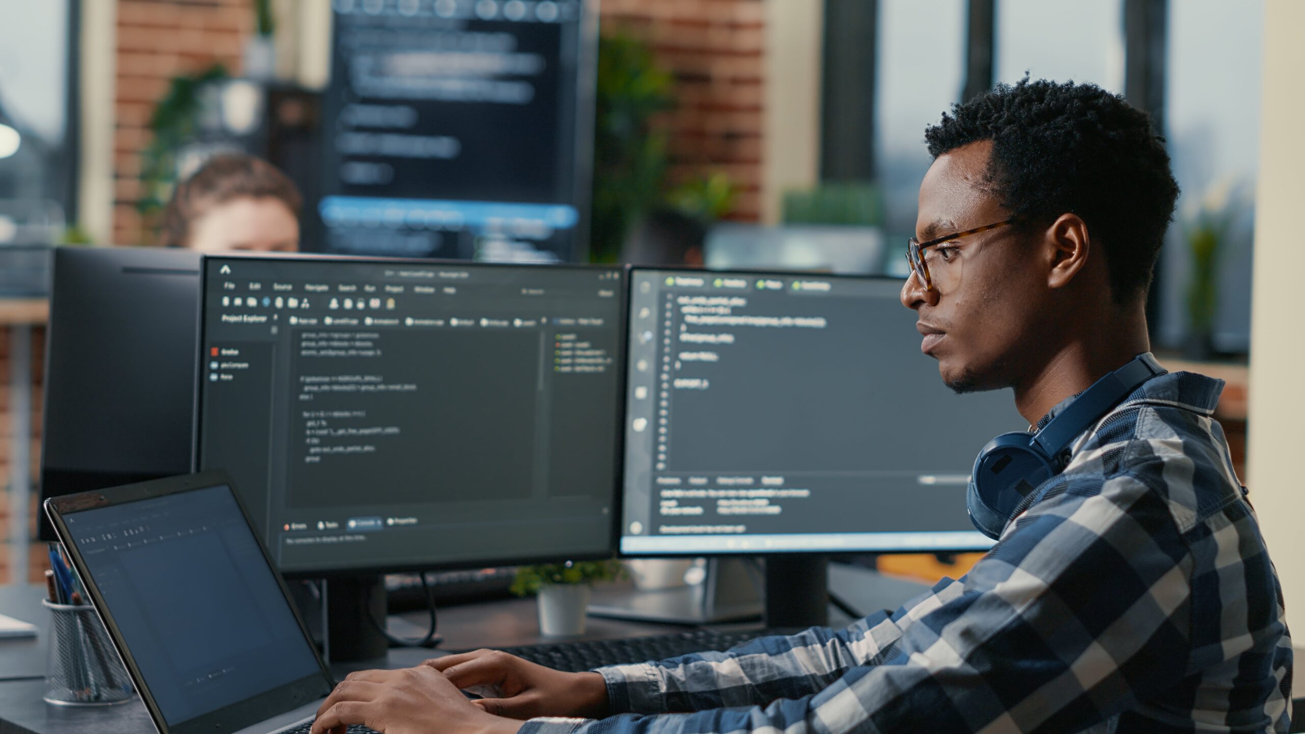 sofware-developer-thinking-while-touching-beard-while-typing-laptop-sitting-desk-with-multiple-screens-parsing-code-focused-database-admin-working-with-team-coding-background-min